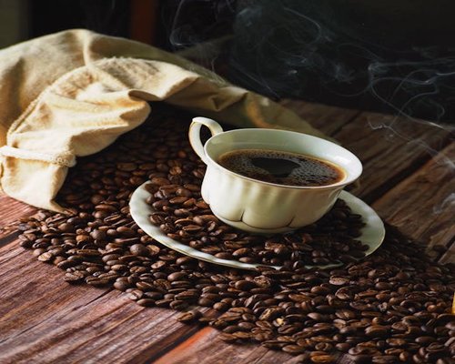 A steaming white ceramic cup of traditional black coffee next to dry coffee beans on a wooden surface